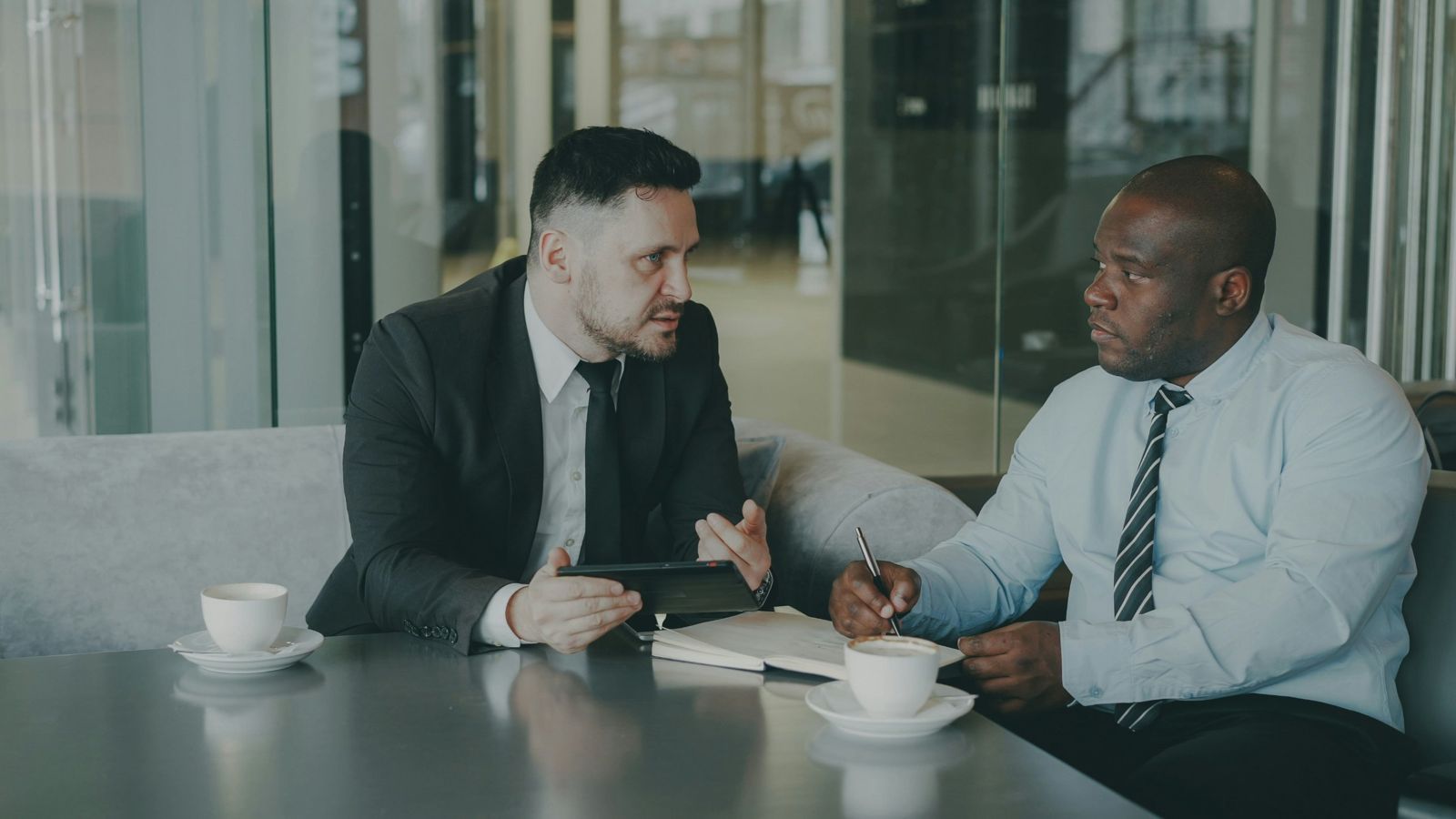 Two men talking over coffee, one taking notes in a notebook